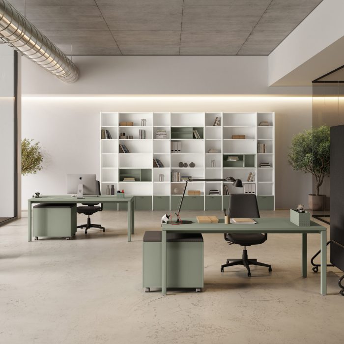 Przestronne biuro w stylu industrialnym z betonowym sufitem i widocznymi rurami wentylacyjnymi. W pomieszczeniu znajdują się dwa minimalistyczne biurka w kolorze szałwiowym, duży biały regał na całą ścianę oraz przeszklona sala spotkań po prawej stronie.