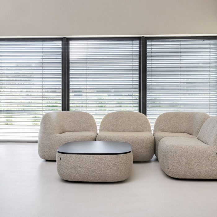 Niska, modułowa sofa o obłych kształtach w kolorze jasnobeżowym, ustawiona w kształcie litery L. Przed nią znajduje się pasujący, niski puf pełniący funkcję stolika kawowego. W tle widać duże okna z żaluzjami.